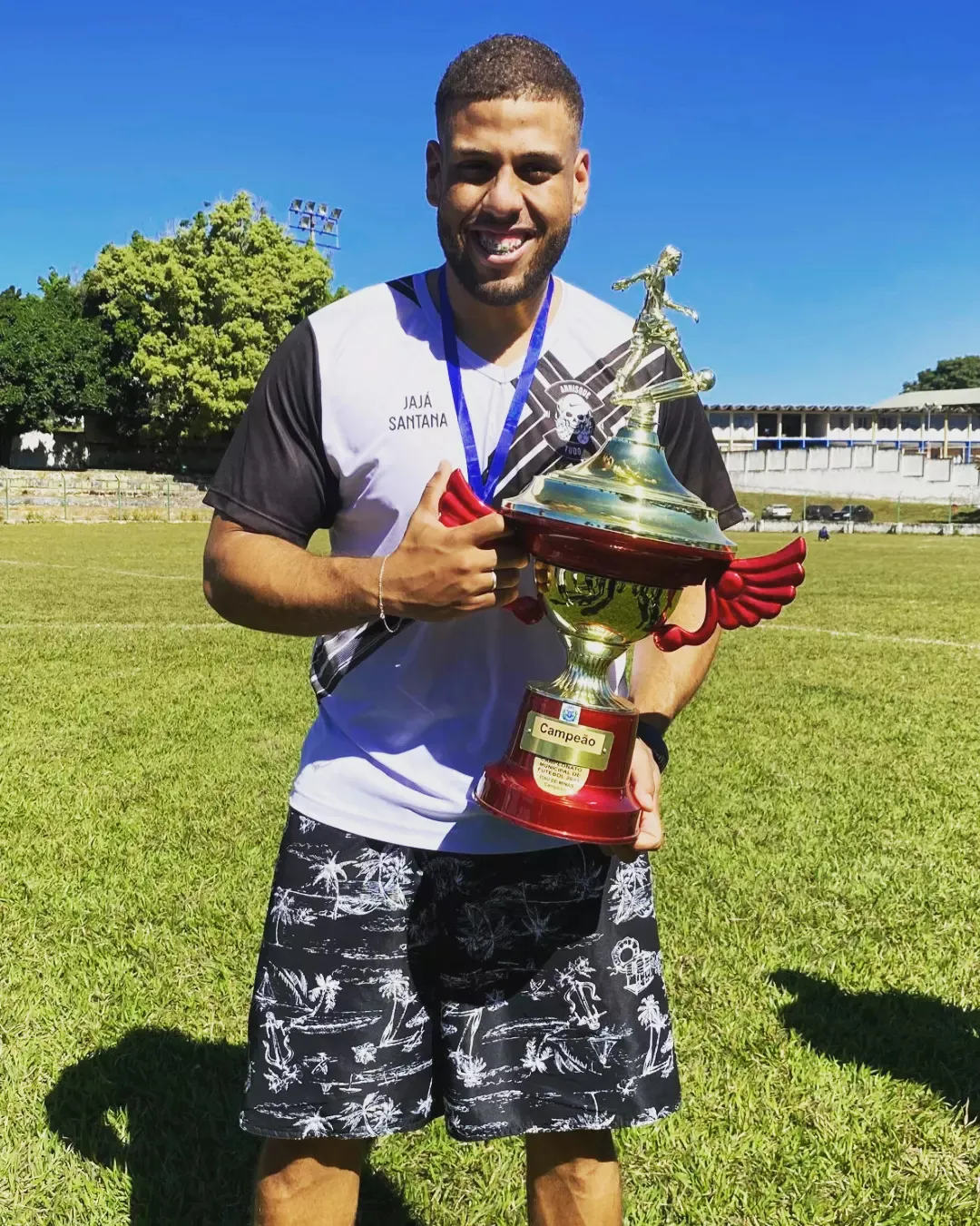 Jajá Santana sorrindo num campo de futebol, de camisa com o nome dele, segurando um troféu de campeão